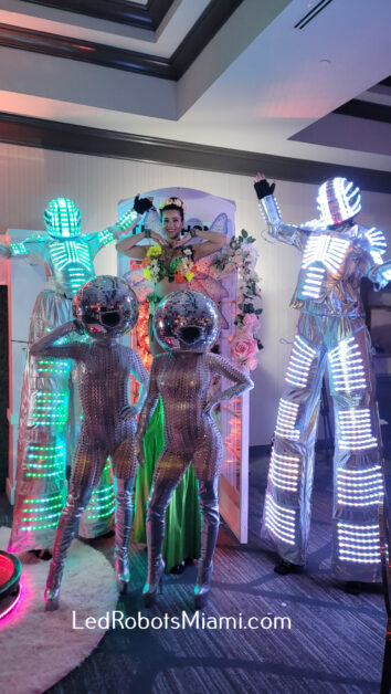 Group of performers in LED costumes, including two tall robots and two smaller reflective figures, pose with a person in a green dress and crown. They're in a bright, colorful setting with floral decorations. Text at the bottom reads "LedRobotsMiami.com". By www.powerparties.com