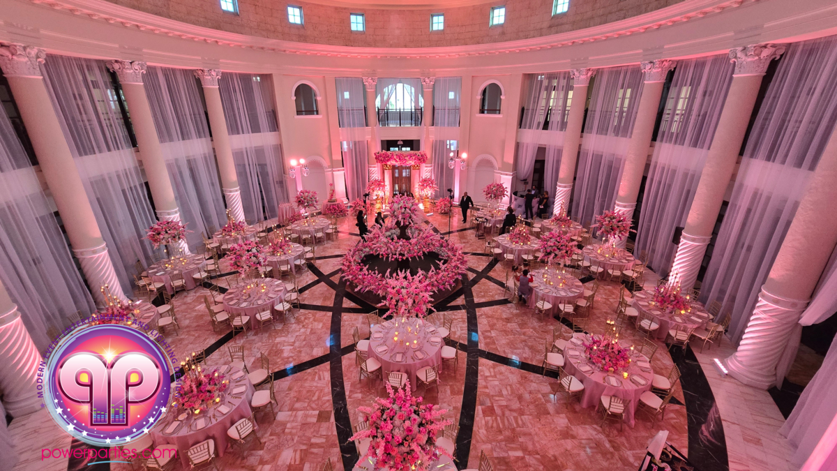 A grand banquet hall beautifully decorated in pink and white for an event. Round tables with floral centerpieces are arranged around a central area, under an elegant dome ceiling. Tall curtains and pillars add to the elegance. A stage is set up at the far end. By www.powerparties.com