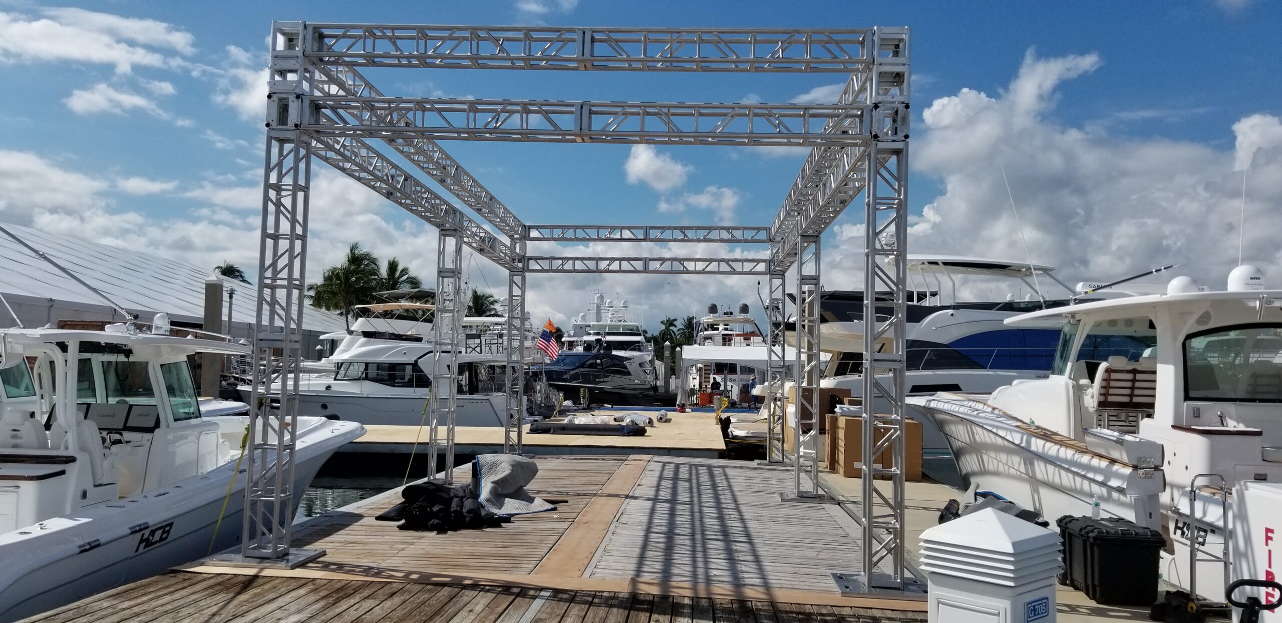 Miami DJ and Hora Locas A marina scene with several docked yachts and a large metallic frame structure in the foreground. The sky is partly cloudy, and there are some people and equipment visible on the dock. By www.powerparties.com