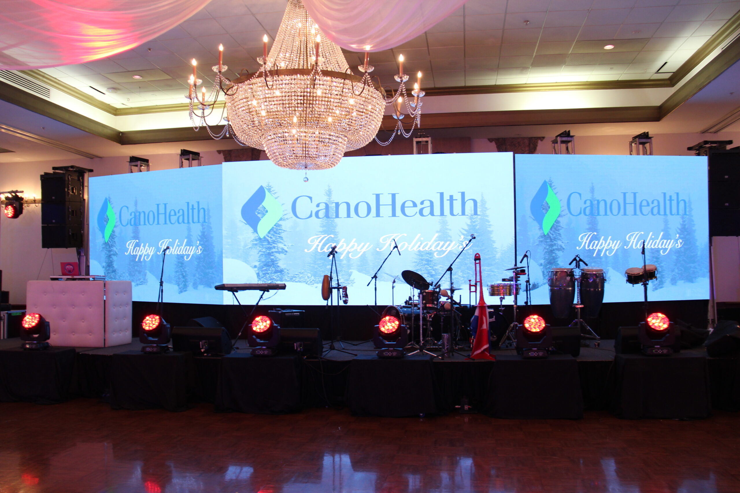 Miami DJ and Hora Locas A decorated event stage for corporate events features three large screens displaying the CanoHealth logo and "Happy Holidays." Musical instruments and various lighting adorn the shiny wooden floor, while a grand chandelier hangs overhead. By www.powerparties.com