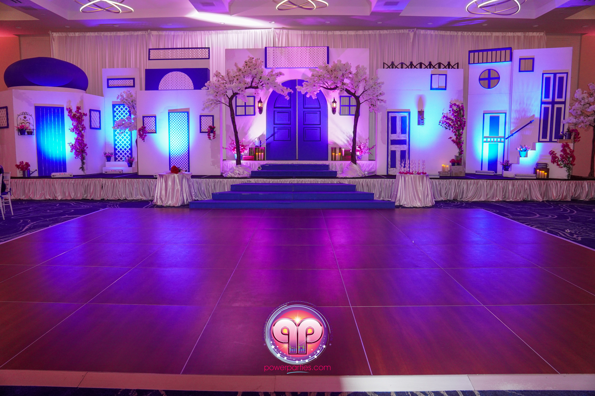 Miami DJ and Hora Locas A vibrant and colorful stage set with a Mediterranean village theme. Blue doors and windows are illuminated with purple and pink lighting. Cherry blossom decorations and candles line the stage. A dance floor is in the foreground. By www.powerparties.com