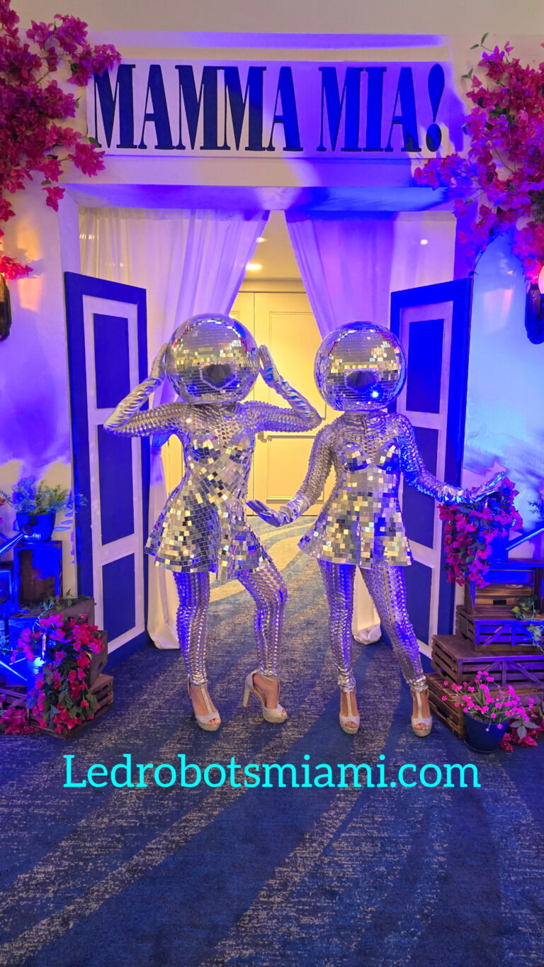 Miami DJ and Hora Locas Two performers in silver sequin bodysuits and mirrored helmets pose in front of an entrance decorated with "MAMMA MIA!" Perfect for a themed quinceañera, they stand against a blue and pink backdrop. The text "Ledrobotsmiami.com" is visible below. By www.powerparties.com