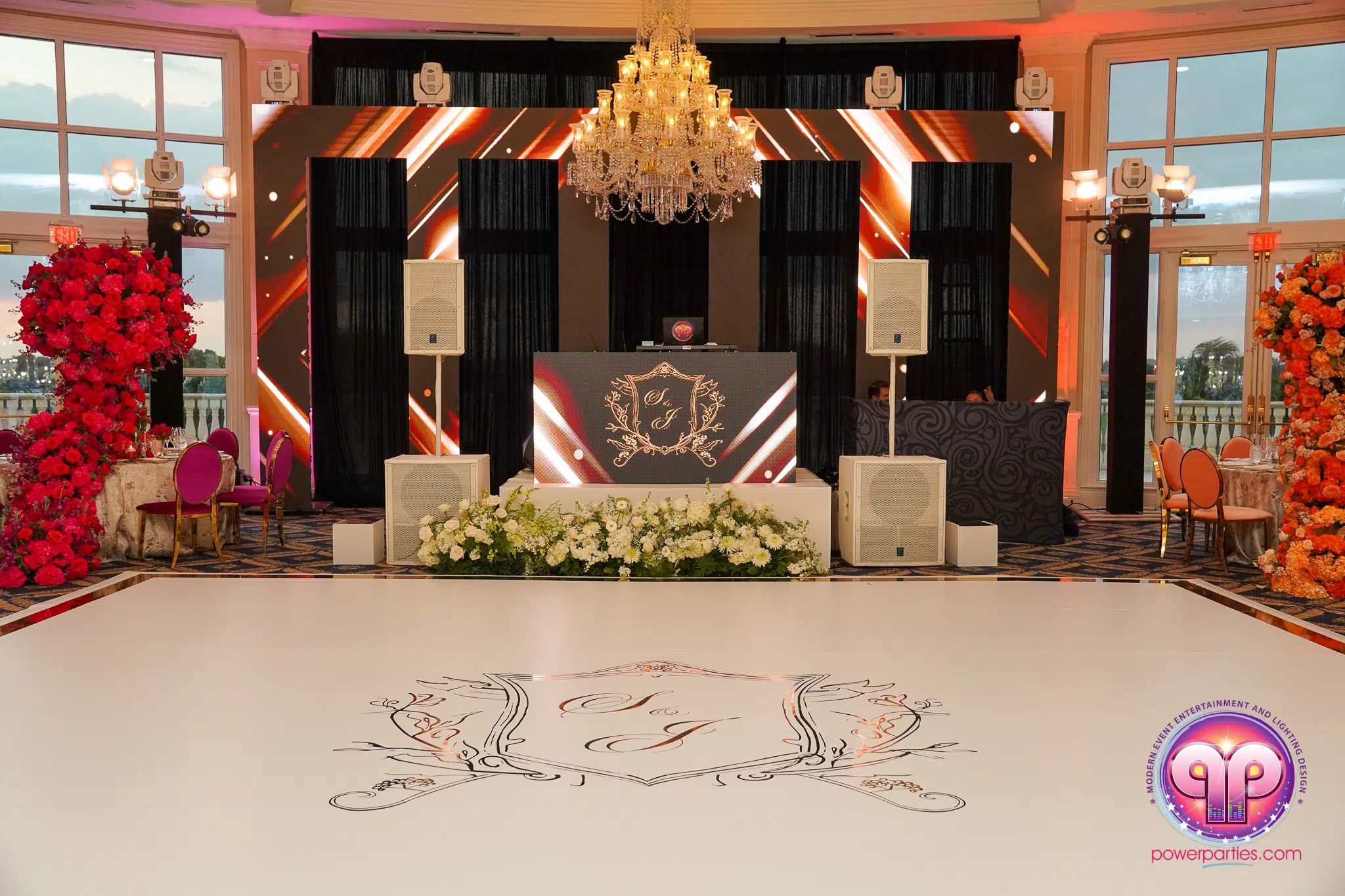 Miami DJ y Hora Locas Elegante montaje de recepción de boda con pista de baile con diseño floral, cabina de DJ con iniciales decorativas, gran lámpara de araña en lo alto, arreglos florales con flores rosas y rojas a ambos lados e iluminación ambiental en un lujoso local. Por www.powerparties.com