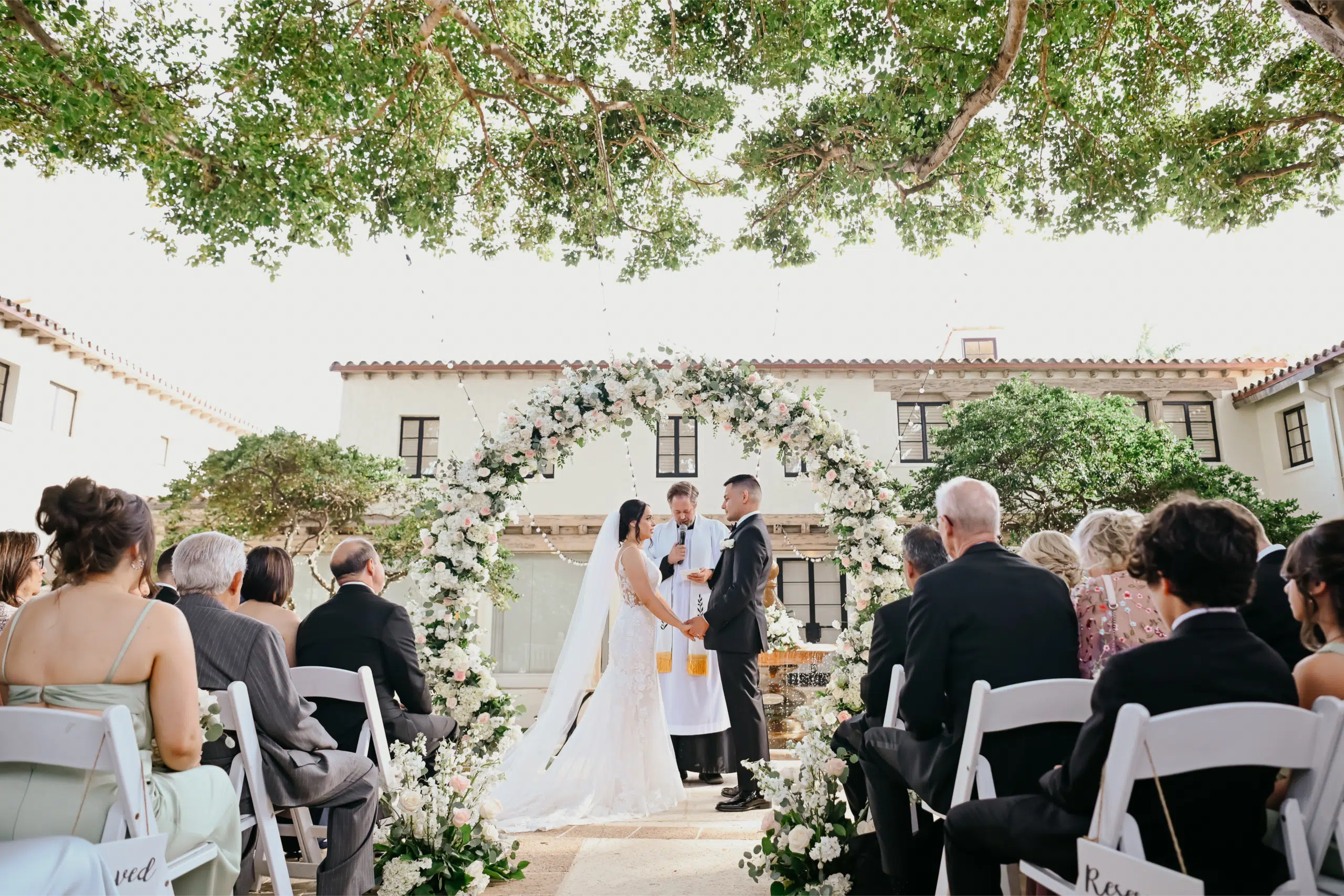 Miami DJ and Hora Locas A couple stands under a large floral arch, holding hands and facing each other during their outdoor wedding ceremony, with guests seated on either side and a building in the background. By www.powerparties.com