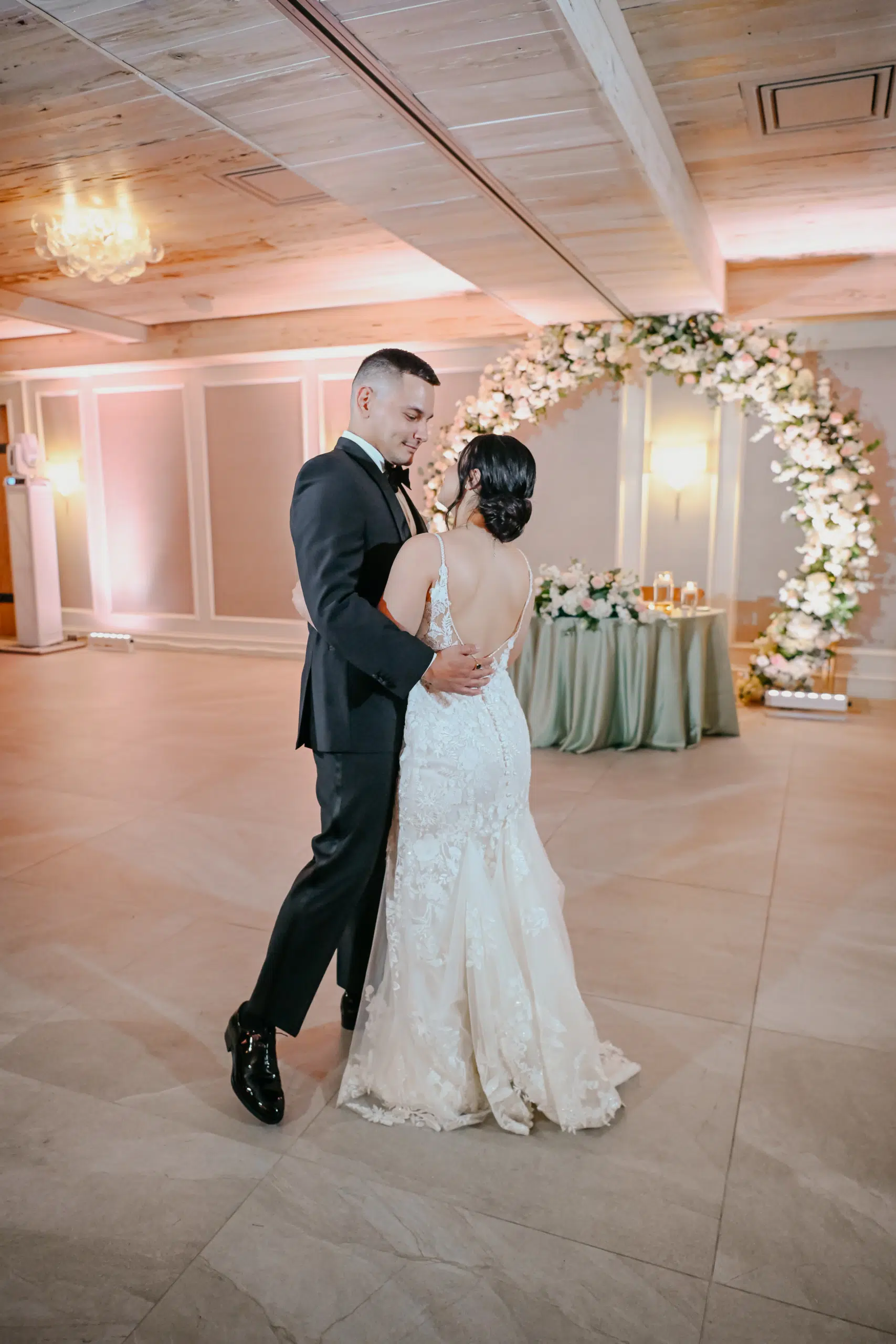 Miami DJ and Hora Locas A bride and groom share their first dance in an elegant, softly lit room with wood ceilings and a floral arch decorated with white and blush flowers in the background. By www.powerparties.com