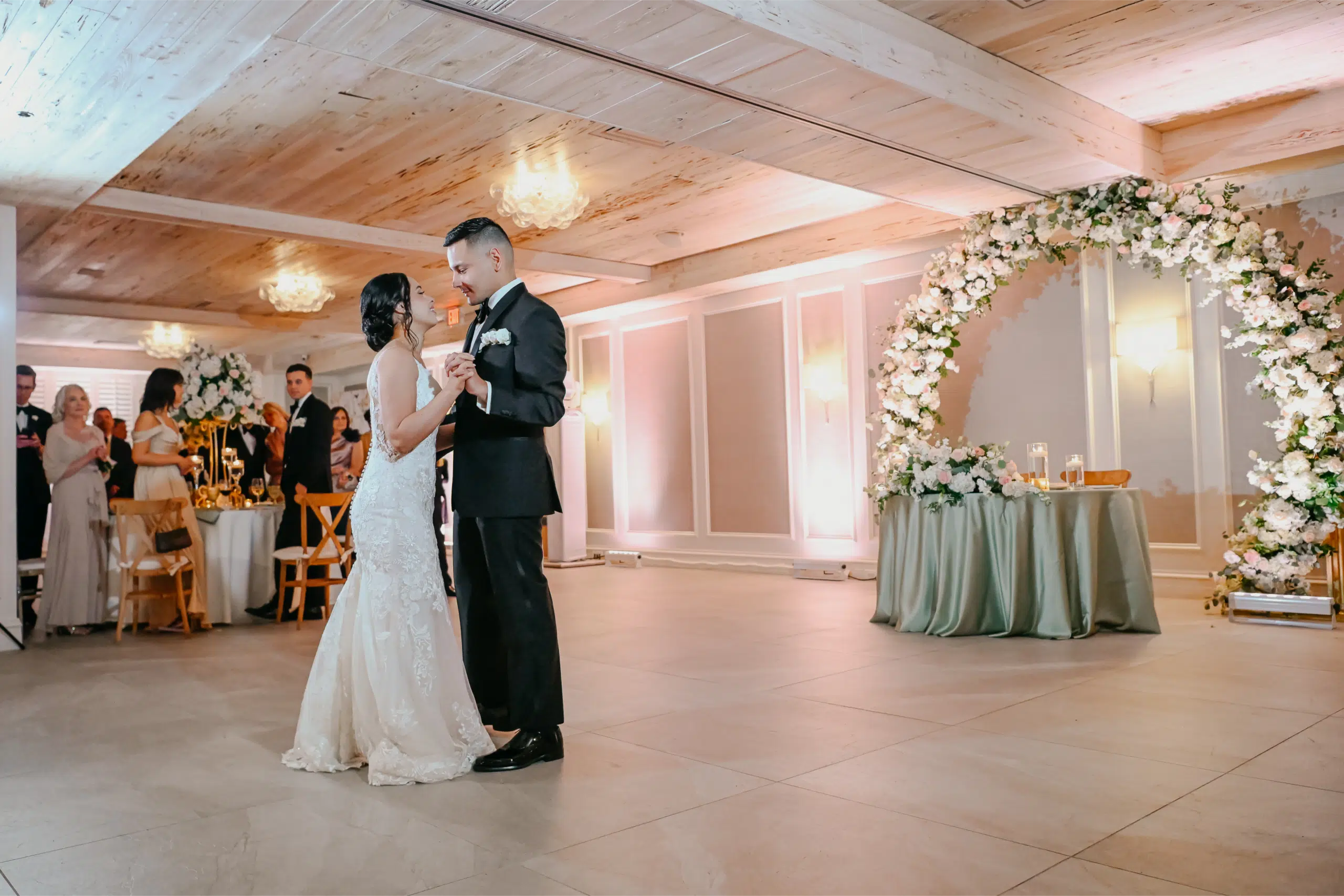 Miami DJ and Hora Locas A bride and groom share their first dance in a warmly lit reception hall, surrounded by guests. A large floral arch and an elegantly decorated table are in the background. By www.powerparties.com