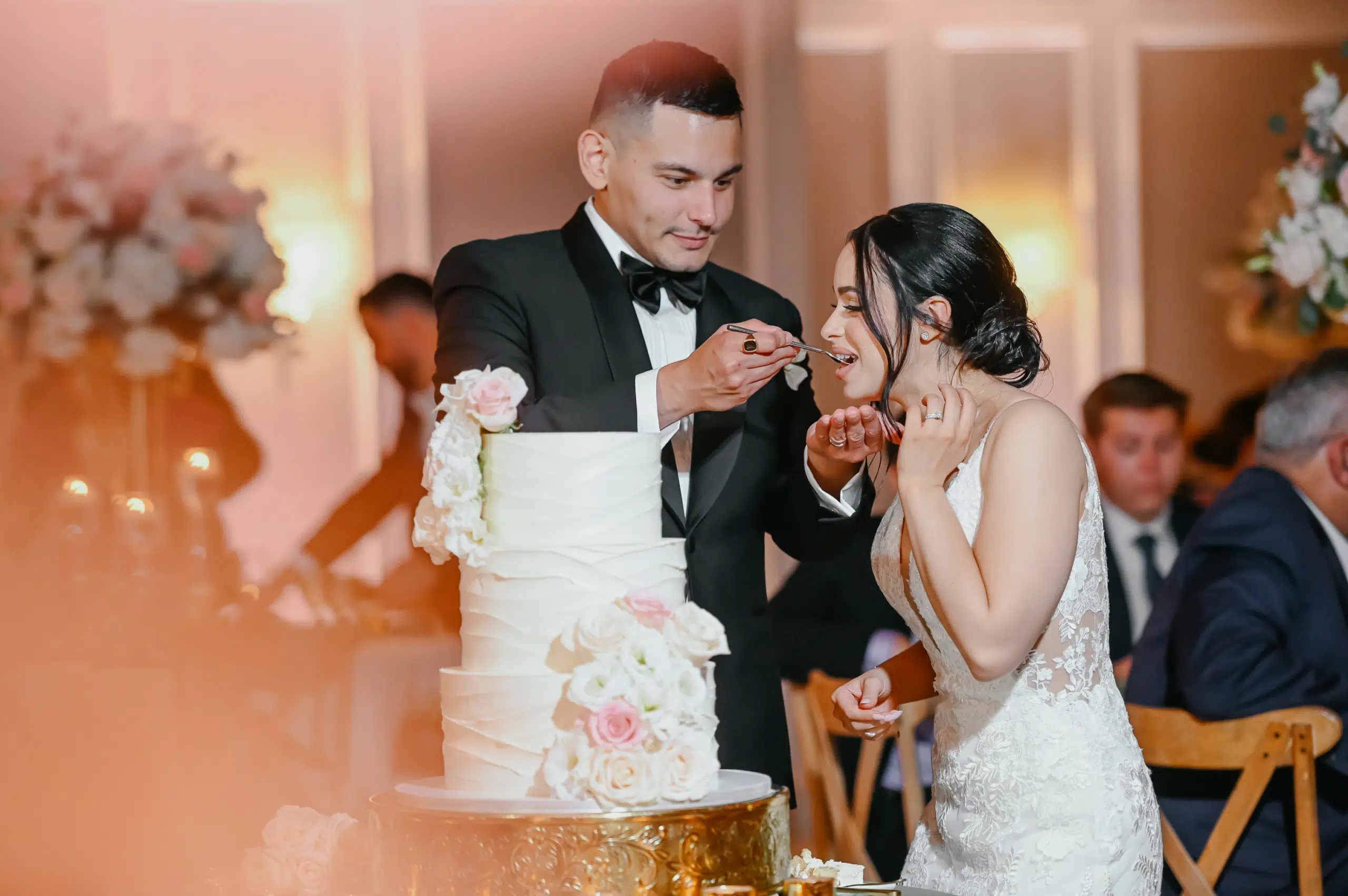 Miami DJ and Hora Locas A groom in a black tuxedo feeds a piece of wedding cake to his bride, who is wearing a white lace dress. The couple smiles, surrounded by floral decorations and seated guests in a warmly lit reception hall. By www.powerparties.com