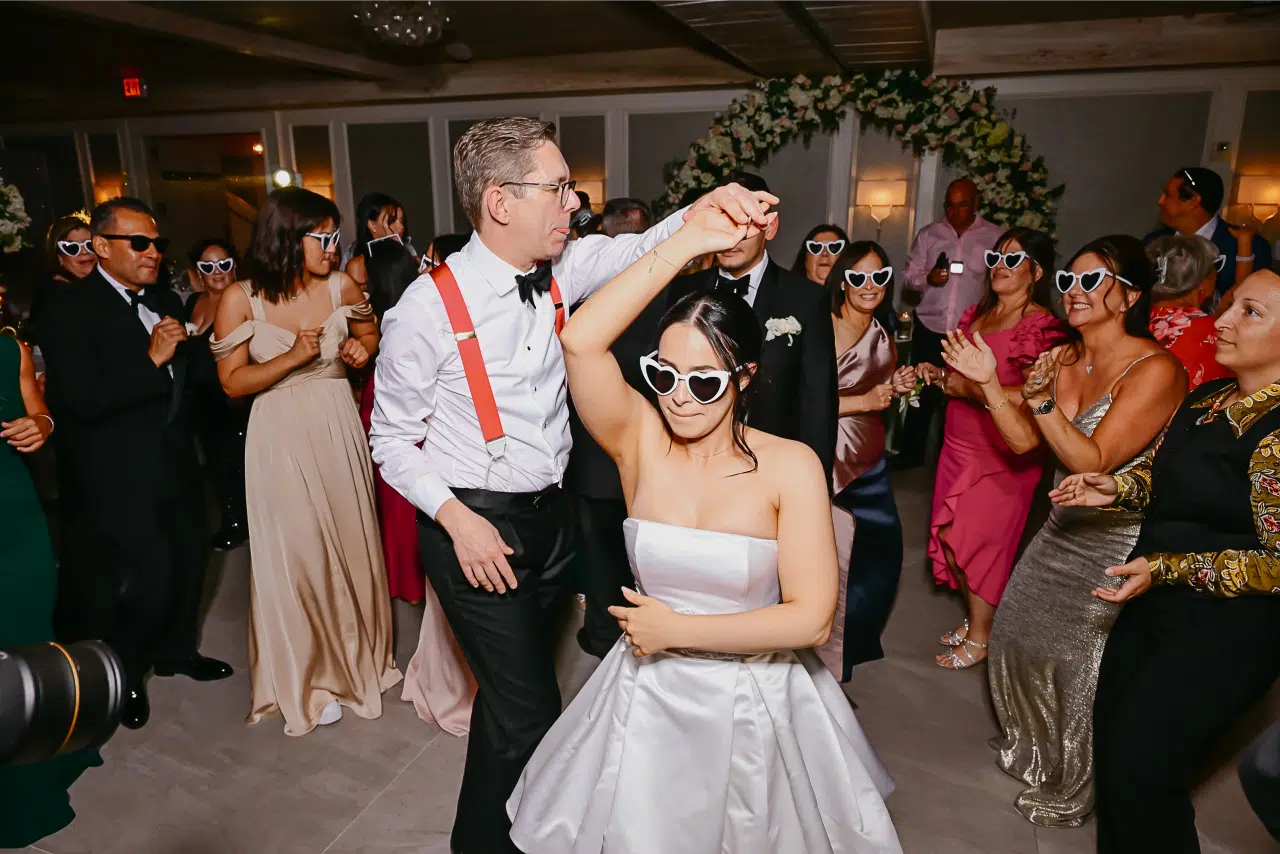 Miami DJ and Hora Locas A bride and groom wearing heart-shaped sunglasses dance joyfully at their wedding reception, surrounded by guests who are also wearing heart-shaped sunglasses and dancing enthusiastically. By www.powerparties.com